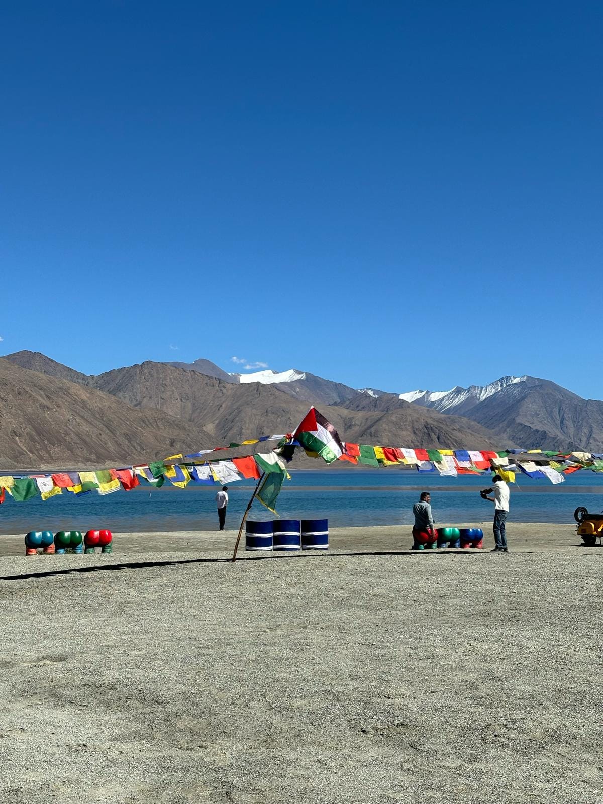 A Palestinian flag is fluttering in the wind in front of a blue lake, surrounded by multicolour prayer flags.