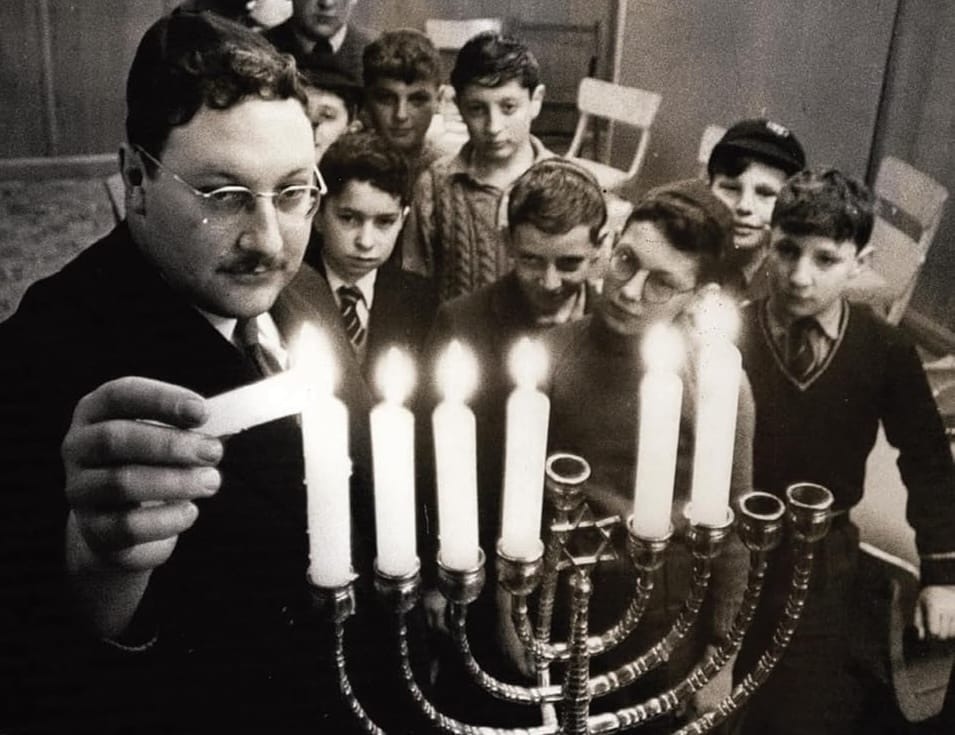 A man is lighting the candles on a menorah. He is surrounded by young boys, some of whom are wearing kippahs. The photo is in black and white.